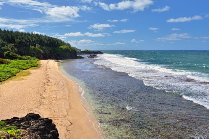 Weitläufiger Sandstrand mit sanften Wellen rechts und üppigen grünen Bäumen links. Der Himmel ist blau mit vereinzelten Wolken, während die Küstenlinie in die Ferne führt.