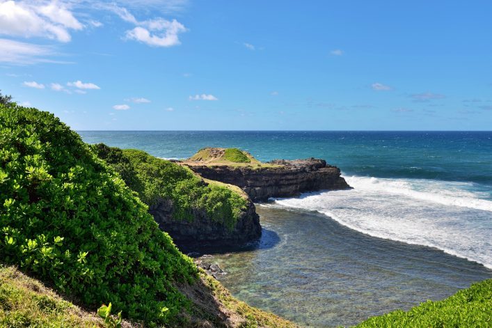 Grüne Küstenklippen erstrecken sich im Vordergrund links, das tiefblaue Meer breitet sich rechts daneben aus. Am Horizont treffen sich das Wasser und der klare, blaue Himmel.