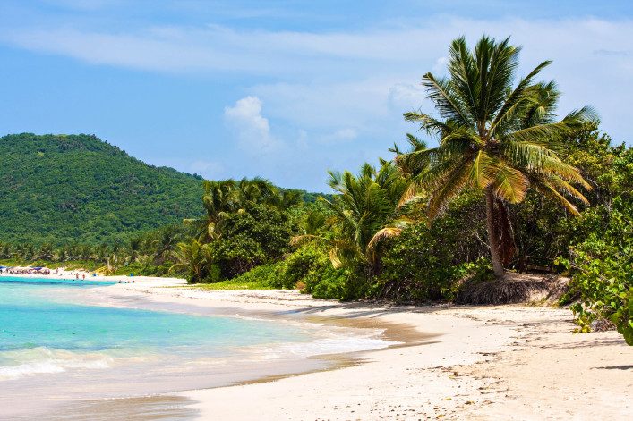 Strand mit weißem Sand, gesäumt von Palmen rechts. Türkisblaues Wasser links und im Hintergrund grüne, bewaldete Hügel. Einige Menschen sind in der Ferne am Strand zu sehen.