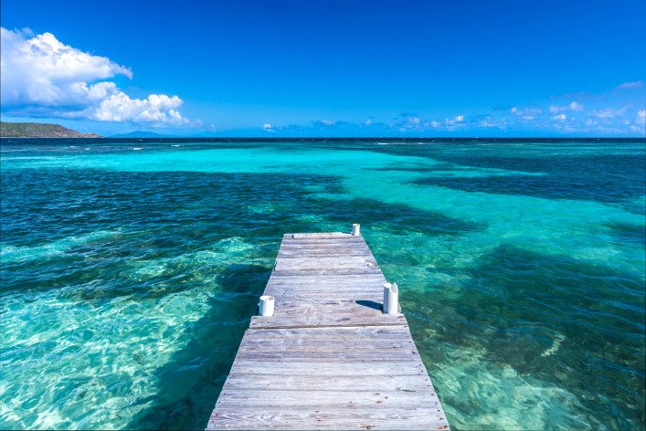 Ein hölzerner Steg führt mittig ins türkisblaue karibische Meer. Links ist eine Hügelküste zu sehen. Der Himmel ist strahlend blau mit ein paar weißen Wolken am Horizont.