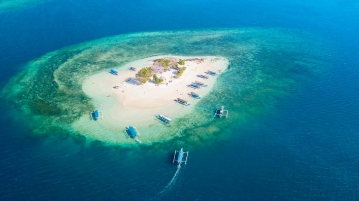 Kleine tropische Sandinsel in klarem blau-grünem Wasser, umgeben von Booten. Auf der Insel sind Palmen und dichtes Grün im Zentrum. Von oben fotografiert, mit Booten um die Insel verteilt.