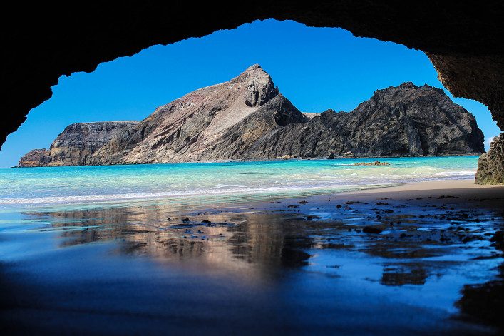 Blick aus einer Höhle auf einen sandigen Strand mit glasklarem Wasser. Im Hintergrund erhebt sich ein felsiger Berg unter strahlend blauem Himmel. Die Höhlenöffnung rahmt die Szene ein.