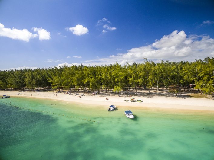 Klarer Himmel mit wenigen Wolken über einem tropischen Strand. Türkisfarbenes Wasser grenzt an den weißen Sandstrand. Boote sind im Wasser verteilt. Im Hintergrund stehen grüne Bäume dicht beieinander.