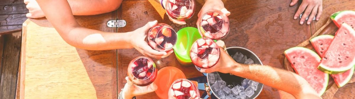 Eine Gruppe feiert ausgelassen mit Sangria am Ballermann auf Mallorca, stoßt an einem sonnigen Tag auf einem Holztisch an, umgeben von Wassermelonenstücken.