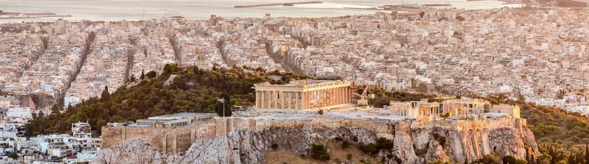 Athen, Akropolis im Sonnenuntergang: Der Parthenon-Tempel thront mittig, umgeben von dicht bebauten Häuserblöcken. Im Hintergrund das glitzernde Meer. Warmes, goldenes Licht über der Szene.