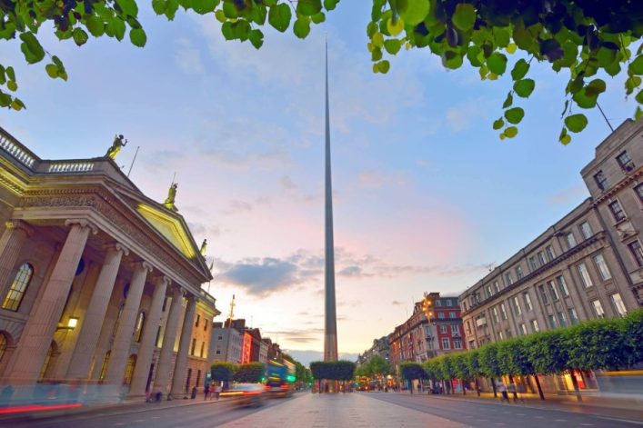 Eine belebte Straße in Dublin bei Dämmerung mit der Spire, einer hohen, schmalen Skulptur, im Zentrum. Links klassizistisches Gebäude, rechts Häuserzeile, grüne Blätter im Vordergrund.