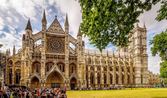 Westminster Abbey mit beeindruckender gotischer Fassade, großen Fenstern und Türmen. Im Vordergrund versammeln sich viele Menschen auf einem Rasen. Rechts und oben Bäume unter blauem Himmel.