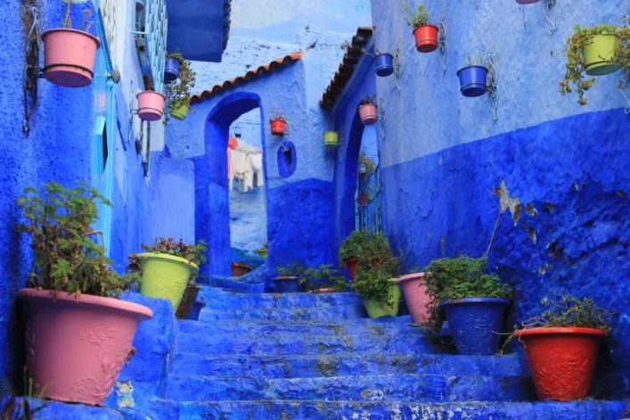 Enge blaue Gasse mit Treppenstufen, gesäumt von bunten Blumentöpfen in rosa, rot und grün. Einige hängen an den Wänden. Ein Torbogen mit Wäsche im Hintergrund. Blauer Himmel sichtbar.