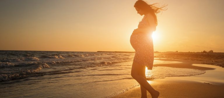 Schwangere Frau genießt ihren Babymoon, steht barfuß am Strand, Umrisse im Sonnenuntergang.