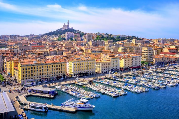 Blick auf den Alten Hafen von Marseille mit Booten und Uferpromenade. Im Hintergrund die Basilika Notre-Dame de la Garde auf einem Hügel.