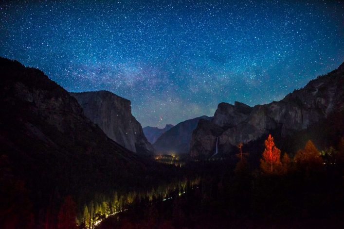 Sternenklare Nacht über dem Yosemite-Tal mit hohen Felsen, darunter der markante El Capitan links und ein Wasserfall rechts. Unten leuchten Bäume im Vordergrund golden.
