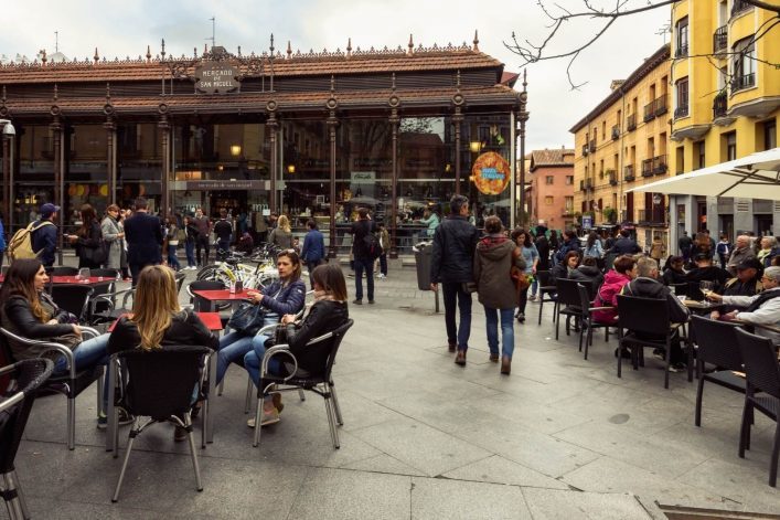 Menschen sitzen an Tischen im Freien vor dem historischen Mercado San Miguel in Madrid. Die Glasfront des Marktes ist umgeben von gelben und rosafarbenen Gebäuden. Belebte Szene mit Freunden.