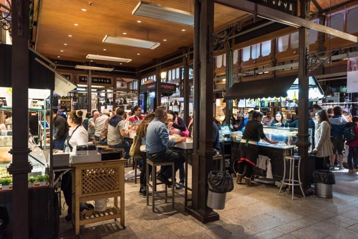 Menschen sitzen und stehen an einem Marktstand im Mercado de San Miguel. Links im Bild bestellen Gäste an einer Theke, während rechts Flaschen über einer Bar hängen. Beleuchteter Holzhimmel.