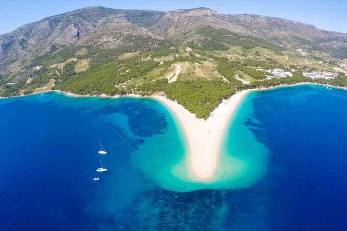 Luftaufnahme des Strandes Zlatni Rat auf der Insel Brac in Kroatien. Eine sandige Landzunge erstreckt sich ins tiefblaue Meer, umgeben von grünen Hügeln im Hintergrund und Booten im Vordergrund.