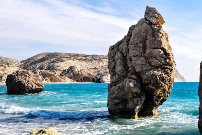Große Felsformation im türkisblauen Meer, umgeben von Wellengang. Im Hintergrund blicken kahle Hügel unter leicht bewölktem Himmel hervor. Der Fels dominiert den Vordergrund rechts.