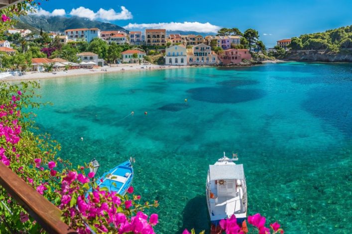 Bunte Häuser an einer Bucht mit türkisem Wasser, umgeben von grünen Bäumen und Blumen, mit Booten im Vordergrund. Bergen im Hintergrund leicht von Wolken umhüllt.