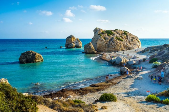 Kleine Bucht mit klarem, blauem Wasser und großen Felsen im Hintergrund. Menschen spazieren am Strand, einige schwimmen im Meer. Vegetation am unteren Bildrand, blauer Himmel mit wenigen Wolken.