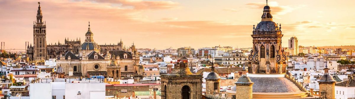 Stadtpanorama von Sevilla bei Sonnenuntergang, im Vordergrund eine verzierte Turmspitze, dahinter historische Gebäude mit Kuppeln und Türmen unter einem orangefarbenen Himmel.
