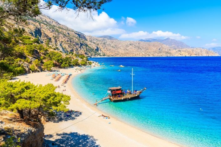 Steile Küstenlinie mit Pinien, türkisfarbenes Meer und Sandstrand. Im Wasser liegt ein Touristenschiff, Sonnenschirme und Liegen am Strand. Perfekter Ort für einen Sommerurlaub oder Strandurlaub.