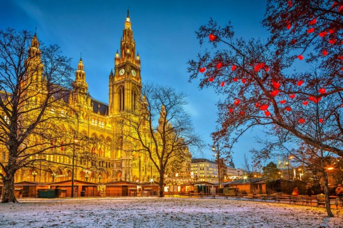 Wiener Rathaus im Winter, prächtig beleuchtet mit goldenen Lichtern. Vor dem Gebäude stehen kahle Bäume, ein Baum rechts ist mit roten Herzlichtern dekoriert. Am Boden liegt eine leichte Schneedecke.