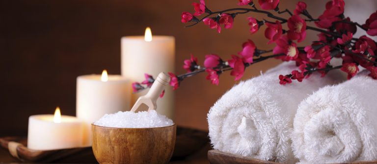 Drei brennende Kerzen stehen links neben einer Schale mit Badesalz auf einem Tisch. Rechts von der Schale befinden sich zwei gerollte Handtücher und rosa Blütenzweige auf einem Holztablett.