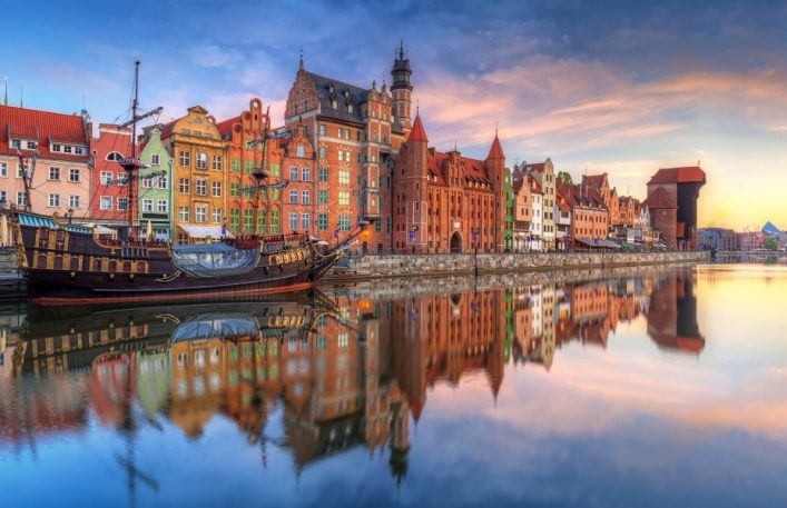 Bunte historische Gebäude an der Motlawa Flussufer in Danzig bei Sonnenuntergang, mit einer Segelschiffsreplik und Spiegelungen im Wasser.