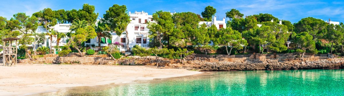 Türkisfarbenes Meer trifft auf feinsandigen Strand, eingerahmt von grüner Vegetation und weißen Häusern – eine idyllische Bucht Mallorcas, ein echter Geheimtipp.