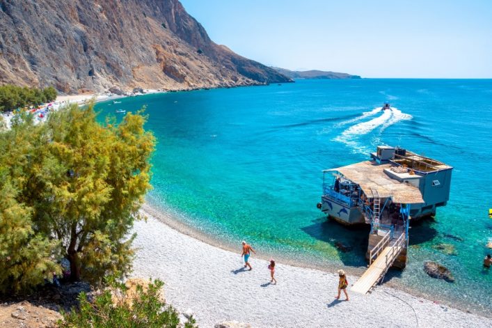Kieselstrand mit türkisblauem Wasser; links Bäume an einer steilen Felsküste, rechts ein schwimmendes Café; Menschen spazieren am Ufer; Motorboot im Hintergrund zieht Spur im Wasser.