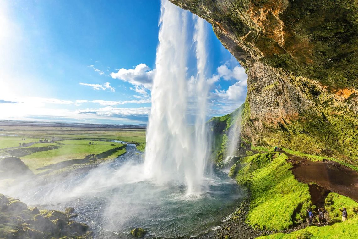 Seljalandsfoss-Wasserfall in Island, stürzt inmitten grüner Landschaft von einer Klippe. Wanderer stehen rechts hinter dem Wasserfall auf einem Pfad. Der Himmel ist blau mit einigen Wolken.