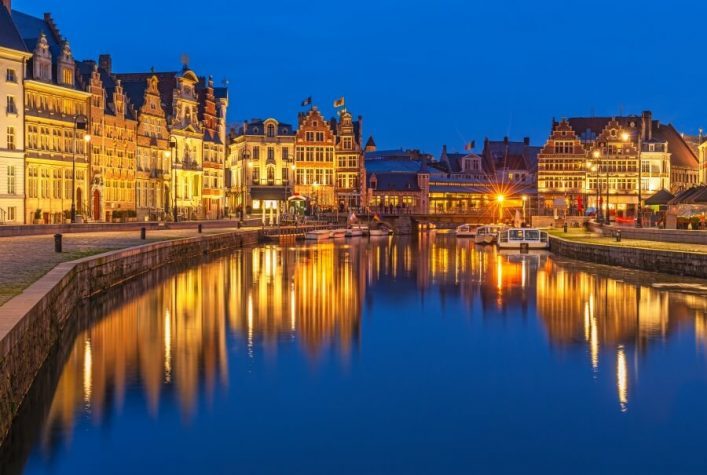 Auf dem Bild sieht man die beleuchtete Stadt Gent bei Nacht. Der Himmel ist dunkelblau und die Stadt spiegelt sich im blauen Wasser wieder.