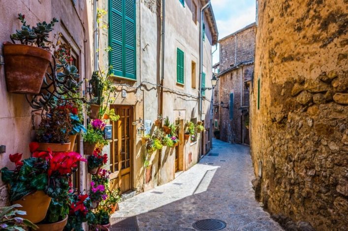 Enge, gepflasterte Gasse in Valldemossa, Mallorca. Links an der Hauswand bunte Blumen in Töpfen, Fensterläden grün. Rechts alte Steinmauer. Klare Himmel des Tages.