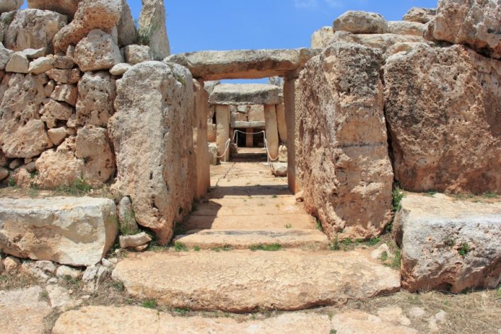 Vor einem blauen Himmel erhebt sich das steinerne Hagar-Qim-Tempelportal in Malta. Massive Kalksteinblöcke stehen links und rechts, ein steiniger Pfad führt durch das Tor ins Innere. Pflanzen sprießen dazwischen.