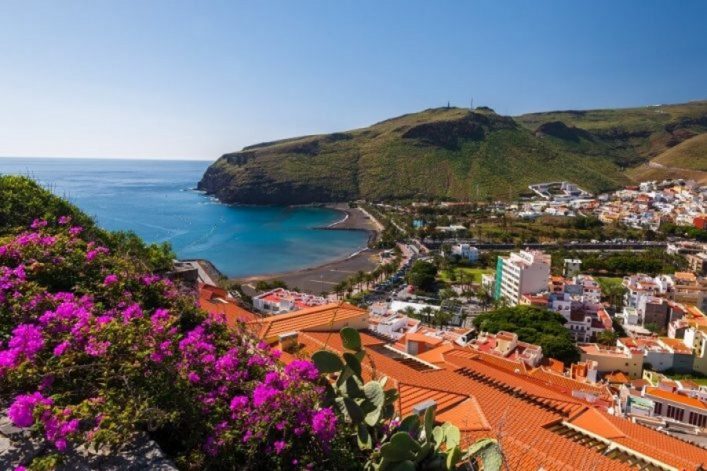 Hügelige Küstenlandschaft mit blauem Meer, palmengesäumtem Strand und bunten Häusern im Hintergrund. Im Vordergrund blühende, pinke Blumen über roten Dächern.