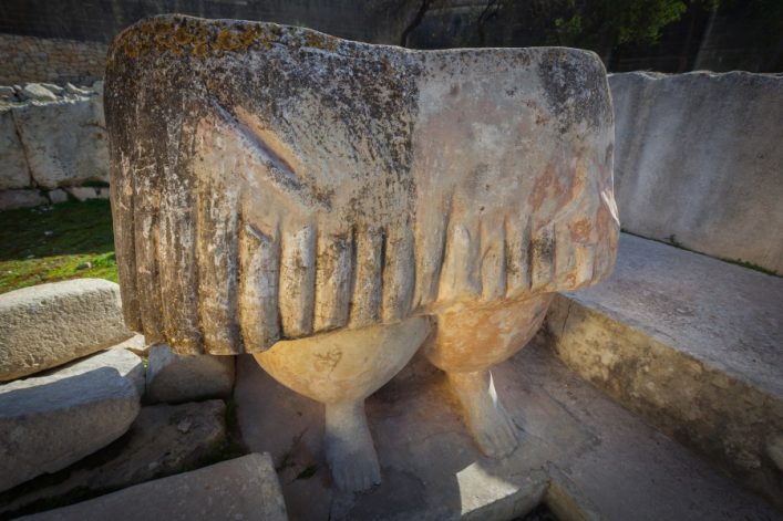Antike Steinfigur mit breiterer Oberseite und zwei beinigem Sockel in einem archäologischen Areal. Im Hintergrund sind verwitterte Mauern und grasbewachsene Flächen zu sehen.
