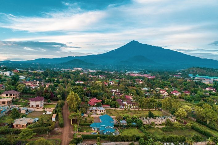 Blick auf Arusha in Tansania mit bunten Häusern, viel Grün und bewaldeten Hügeln im Vordergrund. Im Hintergrund erhebt sich majestätisch der von Wolken umgebene Vulkan Mount Meru.