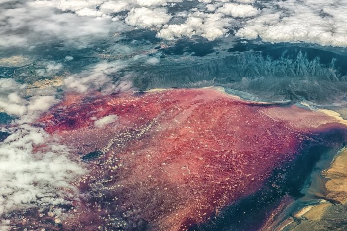 Ein farbenprächtiger See mit tiefrotem und violettem Wasser, umgeben von bewölkten, bergigen Landschaften im Hintergrund und wenigen weißen Wolken im Vordergrund. Die Farben des Wassers verlaufen sanft ineinander.