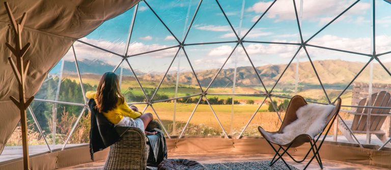 Eine Person sitzt in einem Korbstuhl in einem geodätischen Zelt. Große Panoramafenster bieten eine beeindruckende Aussicht auf eine hügelige Landschaft. Im Zelt steht noch ein Stuhl mit Fellbezug.