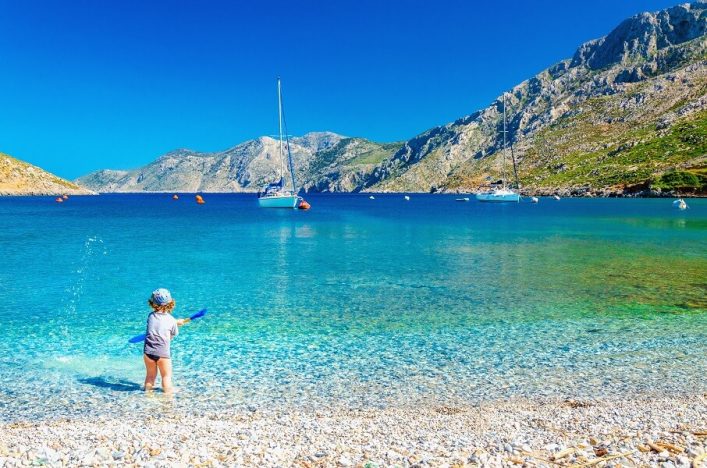 Am Kiesstrand spielt ein Kind im flachen, klaren Wasser. Im Hintergrund segeln zwei Boote auf dem türkisblauen Meer, umgeben von grünen Bergen unter strahlend blauem Himmel.