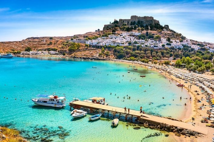 Strand mit türkisem Wasser; Menschen schwimmen und entspannen. Rechts verläuft ein vollbesetzter Sandstrand mit Sonnenschirmen. Im Hintergrund ein Hügel mit weißen Häusern und einer antiken Festung.