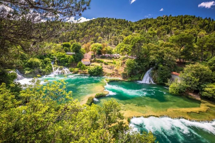 Grüne Baumlandschaft umrahmt türkisfarbenen Wasserfall in Skradinski Buk, Krka-Nationalpark, Kroatien. Wasser fließt in Becken zwischen grünen Inseln. Ein Steinhaus und Wald bedecken den Hintergrund.