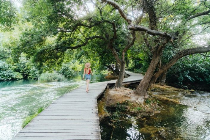 Eine Frau mit Sonnenhut und rotem Tanktop spaziert auf einem Holzsteg durch den üppig grünen Krka Nationalpark. Links fließt ein klarer Fluss, rechts stehen knorrige Bäume.