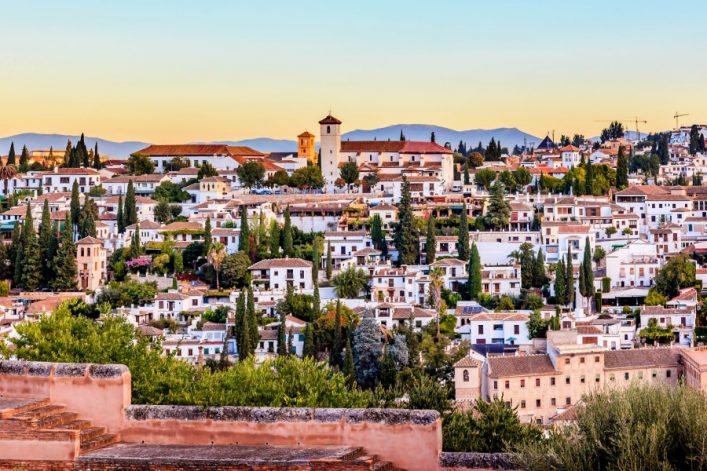 Im Vordergrund Ziegeldächer und grüne Bäume; im Hintergrund die Alhambra mit beige-roter Fassade. Dahinter Berge unter blauem Himmel in der Abenddämmerung.