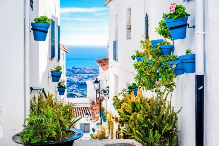 Pflanzen in blauen Töpfen hängen an weißen Hauswänden, eine schmale Gasse führt bergab zum Meer. Auf beiden Seiten stehen grüne Pflanzen, im Hintergrund erstreckt sich das azurblaue Meer.