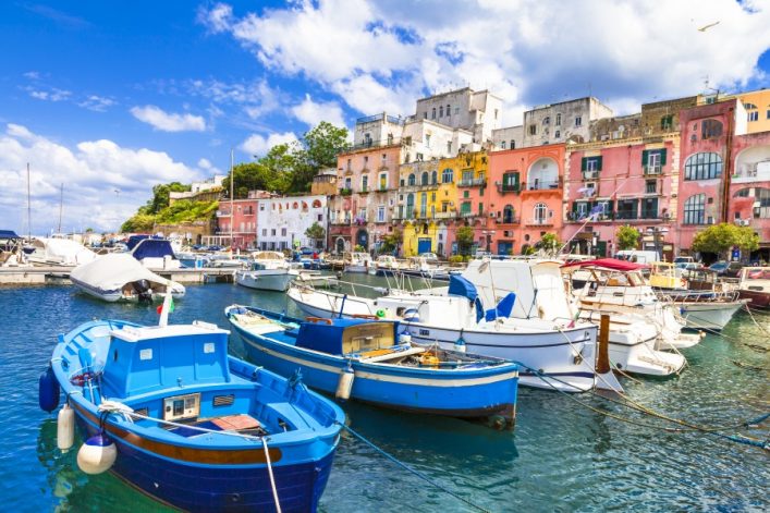 Bunte Häuser reihen sich entlang eines Hafenbeckens auf Procida, Italien. Im Vordergrund ankern blaue und weiße Boote auf ruhigem Wasser unter blauem Himmel.