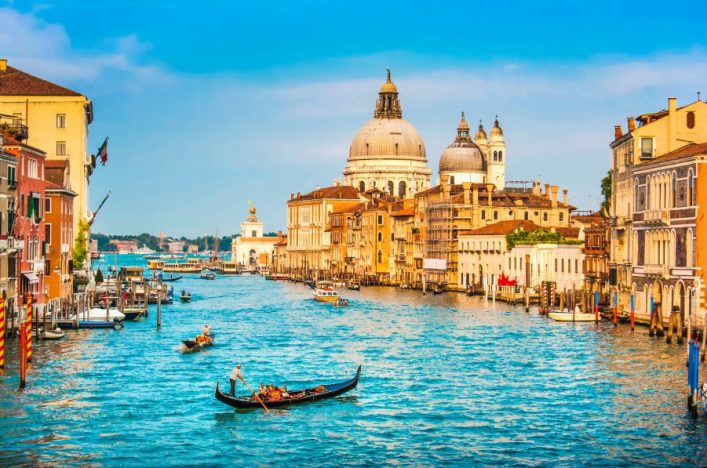 Gondel auf dem Canal Grande in Venedig, umgeben von historischen Gebäuden links und rechts. Im Hintergrund erhebt sich eine große Kuppelkirche unter klarem, blauem Himmel.