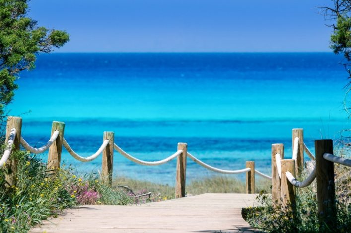 Holzweg mit Seilgeländern führt durch grüne Vegetation zum türkisfarbenen Meer unter klarem, blauem Himmel. Bäume rahmen den Weg links und rechts ein, Blumen blühen am Wegesrand.