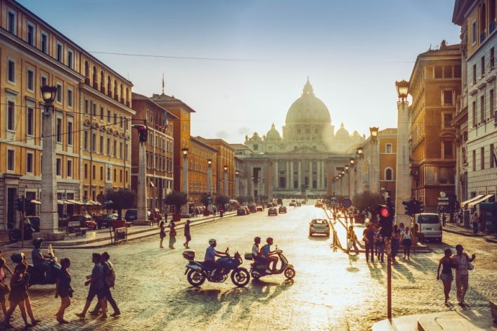 Breite Straße in Rom bei Sonnenuntergang, flankiert von historischen Gebäuden. Menschen spazieren oder fahren Roller. Im Hintergrund erhebt sich eindrucksvoll eine große Kuppel.