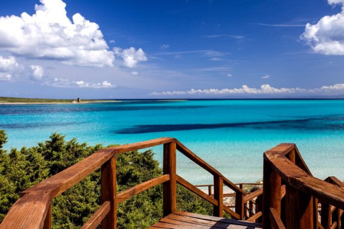 Holzsteg führt durch grüne Büsche zum strahlend türkisfarbenen Meer; am Horizont kleine Insel und Wolken am blauen Himmel; links im Hintergrund alter Turm.