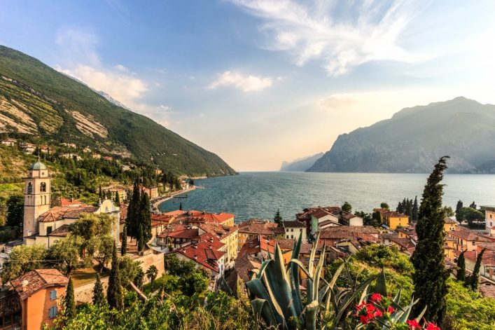 Blick auf Torbole am Gardasee mit roten Ziegeldächern, grünen Zypressen und einer Kirche links im Vordergrund. Der blaue See wird von hohen Bergen eingerahmt, der Himmel ist leicht bewölkt.