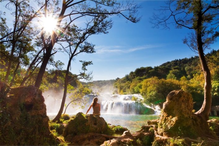 Eine Frau sitzt auf einem Felsen, umgeben von Bäumen, und blickt auf einen beeindruckenden Wasserfall im Krka-Nationalpark. Die Sonne scheint durch die Baumkronen und taucht die Szene in ein warmes Licht